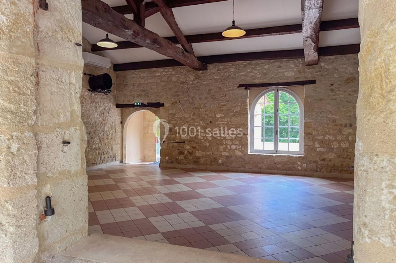 Salle en pierre avec poutres apparentes, carrelage au sol, grande fenêtre et porte voûtée ouverte sur l'extérieur.