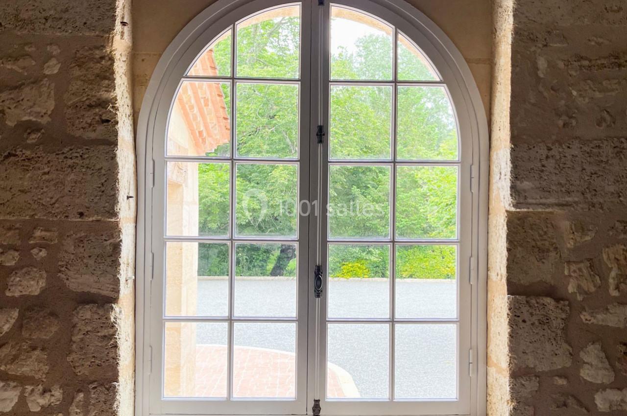 Fenêtre cintrée en bois blanc donnant sur un jardin verdoyant, encadrée par un mur en pierre beige.