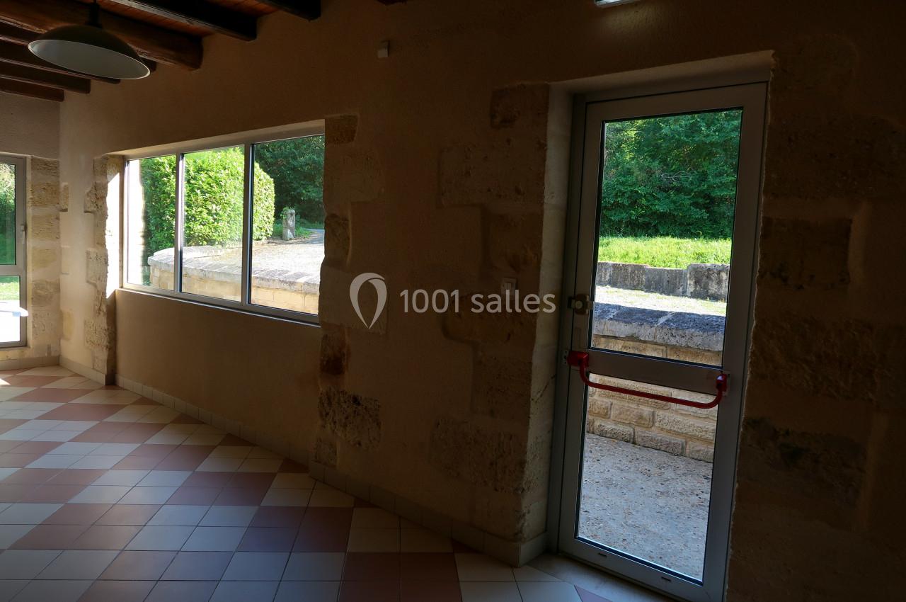 Vue intérieure d'une pièce avec murs en pierre, carrelage au sol, fenêtre et porte vitrée donnant sur un espace extérieur…