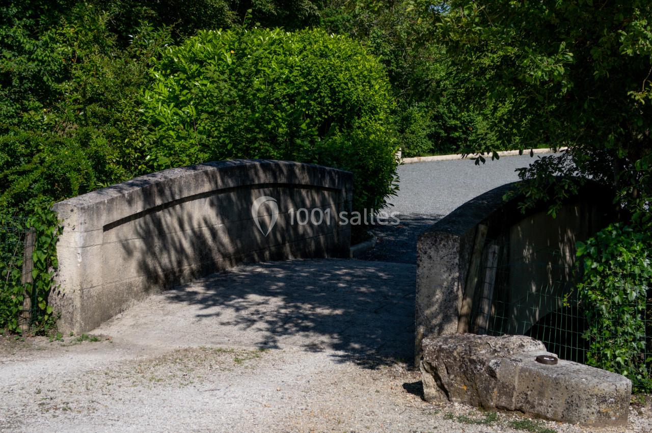Petit pont en pierre entouré de végétation, menant à un chemin gravillonné sous un ciel dégagé.