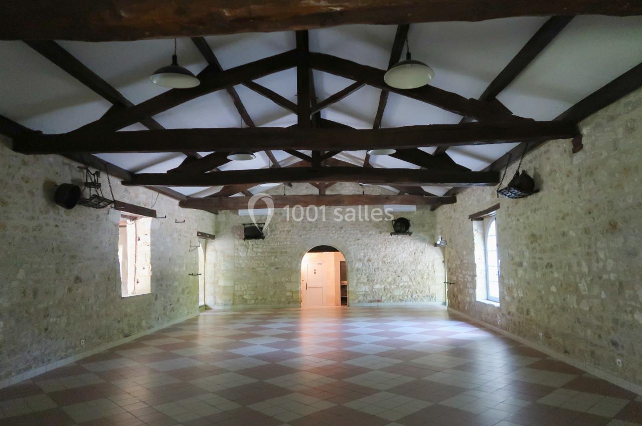 Salle intérieure en pierre avec poutres apparentes, sol carrelé et lumière naturelle entrant par des fenêtres.