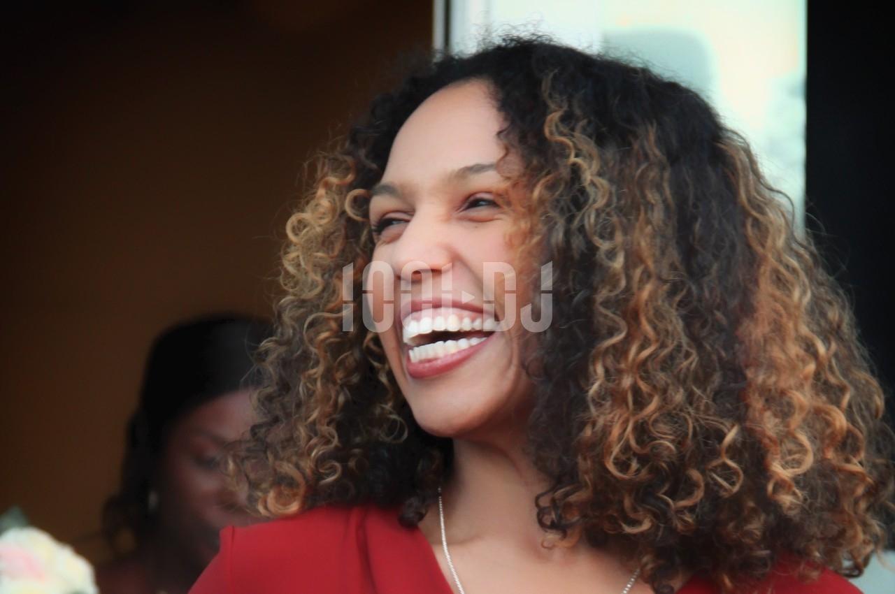 Femme souriante aux cheveux bouclés, portant un haut rouge, photographiée en extérieur avec un fond flou.