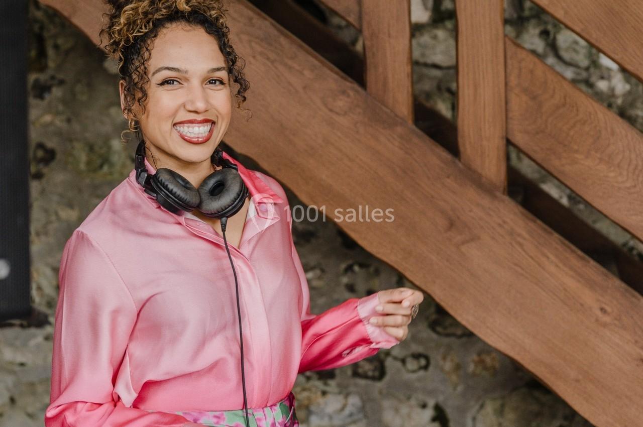Femme souriante portant un casque audio autour du cou, debout devant un escalier en bois et un mur en pierre.