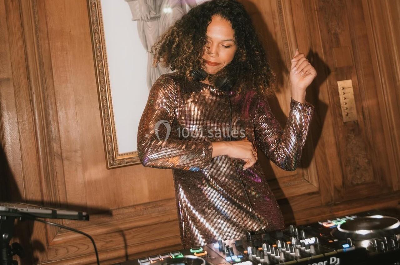 Une femme en robe scintillante mixe de la musique sur une table de DJ dans une pièce aux murs en bois.