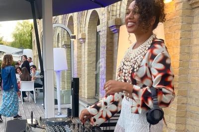 Une DJ souriante mixe en extérieur sur une terrasse, entourée de convives assis et debout.