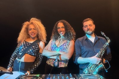 Trois musiciens posent sur scène avec une table de mixage et un saxophone, éclairés par des projecteurs.