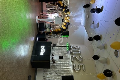 Salle décorée pour une fête avec ballons noirs et dorés, chiffres ’2025’ et une table de DJ en avant-plan.