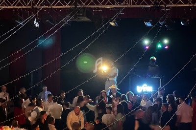 Salle de réception animée avec des invités dînant et dansant sous une lumière tamisée, DJ en arrière-plan.