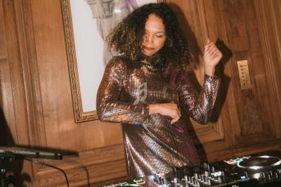 Une femme en robe scintillante mixe de la musique sur une table de DJ dans une pièce au décor classique en bois.