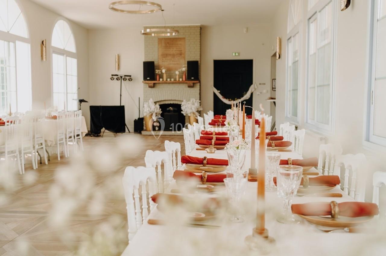 Salle de réception lumineuse avec tables décorées de nappes blanches, serviettes rouges et bougies.