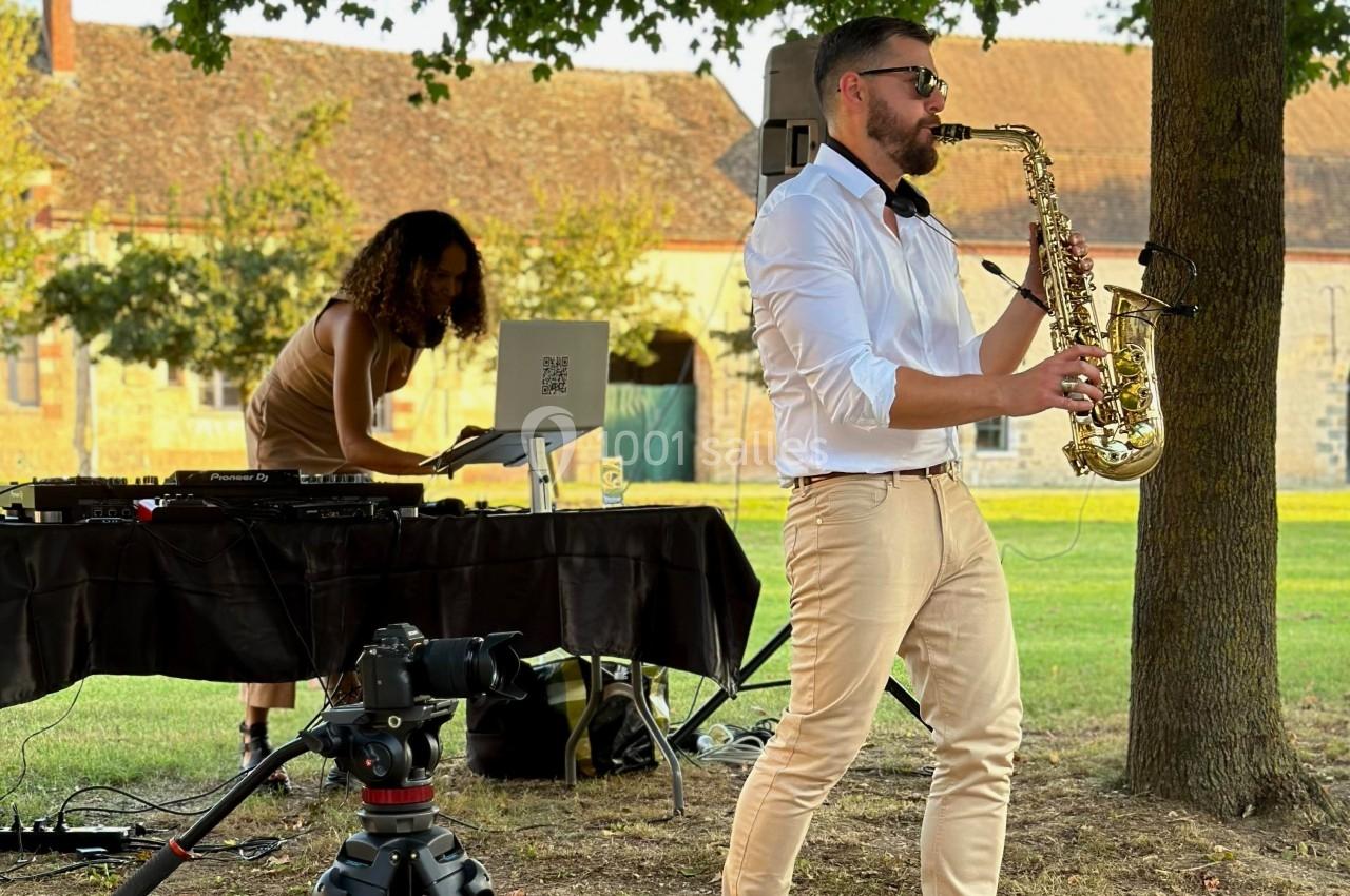 Un saxophoniste joue en plein air près d'une DJ animant une table de mixage, dans un cadre verdoyant.