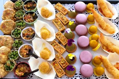 Table dressée en extérieur avec des plats, ustensiles de cuisine et présentoirs en bois devant une haie verte.