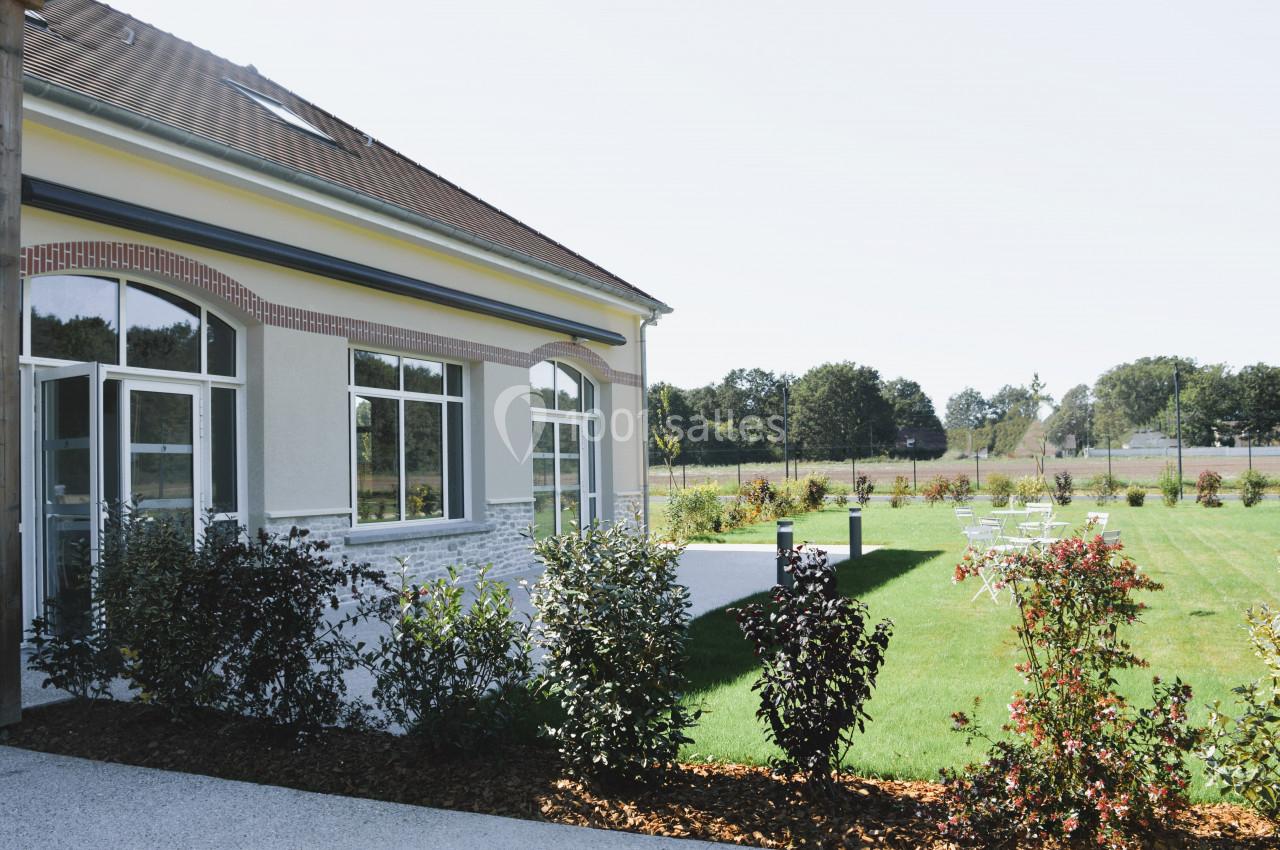 Bâtiment avec de grandes fenêtres donnant sur un jardin aménagé avec pelouse, arbustes et quelques chaises blanches.