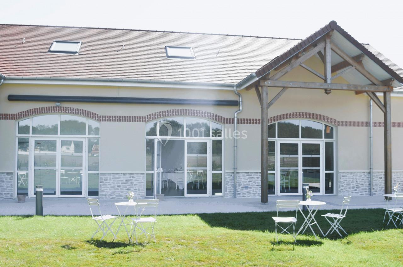 Façade d'un bâtiment moderne avec de grandes baies vitrées, terrasse aménagée avec tables et chaises sur pelouse.