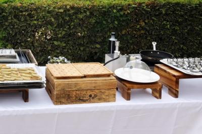 Table dressée en extérieur avec des plats, ustensiles de cuisine et présentoirs en bois devant une haie verte.