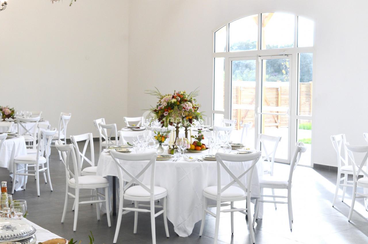 Salle de réception lumineuse avec tables rondes dressées, décorations florales et vaisselle disposée au premier plan.