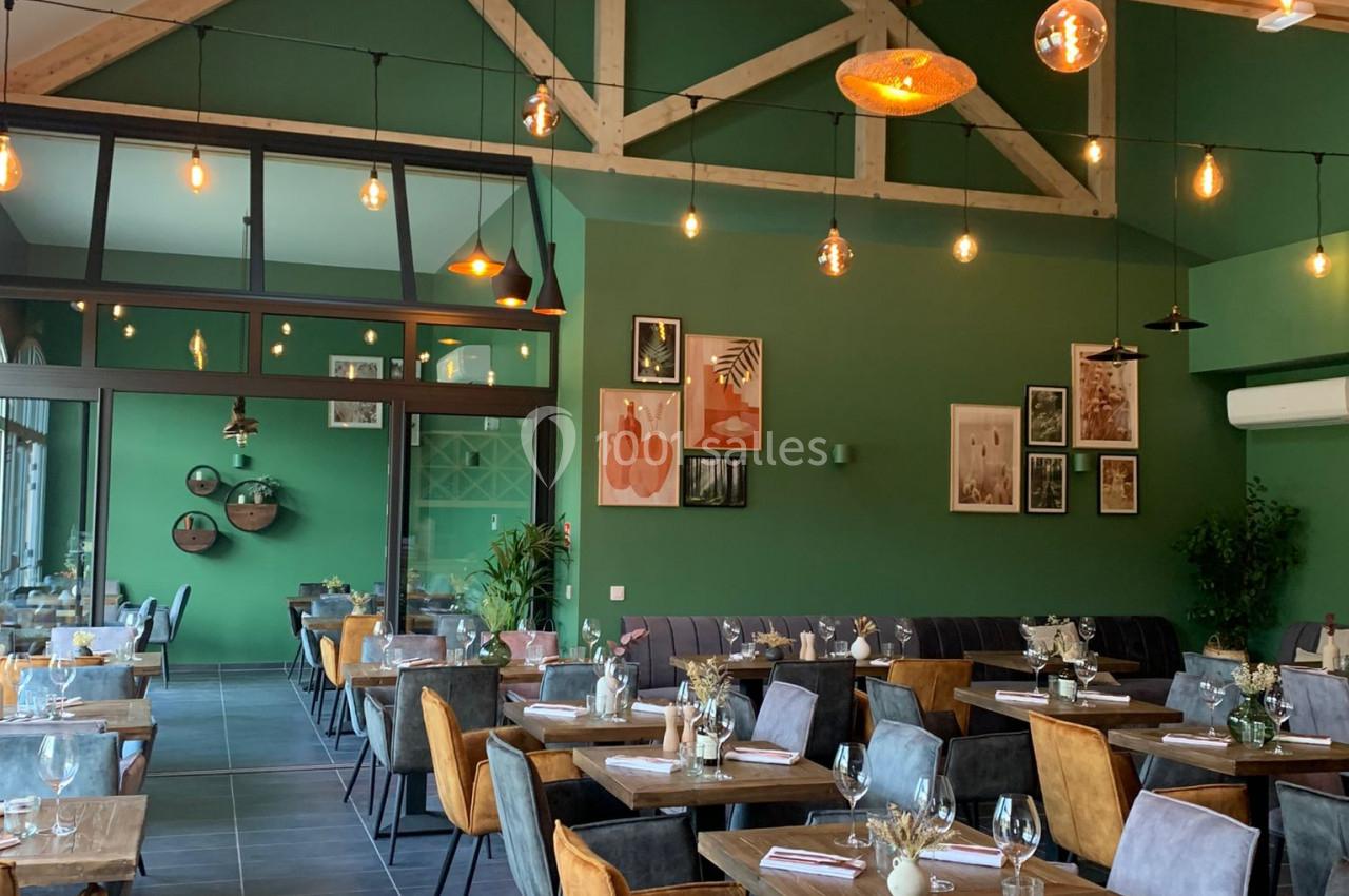 Salle de restaurant moderne avec murs verts, poutres apparentes, tables dressées et éclairage suspendu chaleureux.