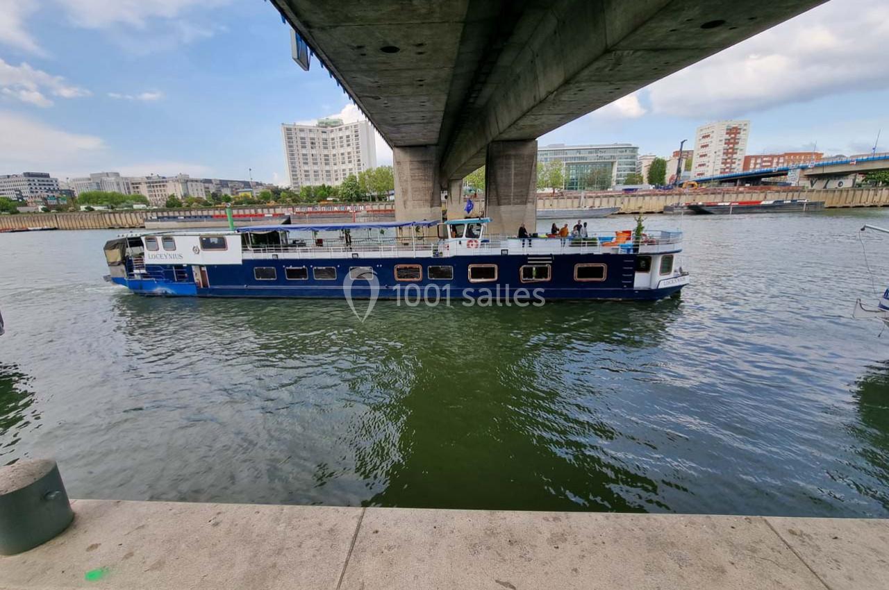 Bateau bleu naviguant sous un pont en milieu urbain, avec des bâtiments modernes en arrière-plan.