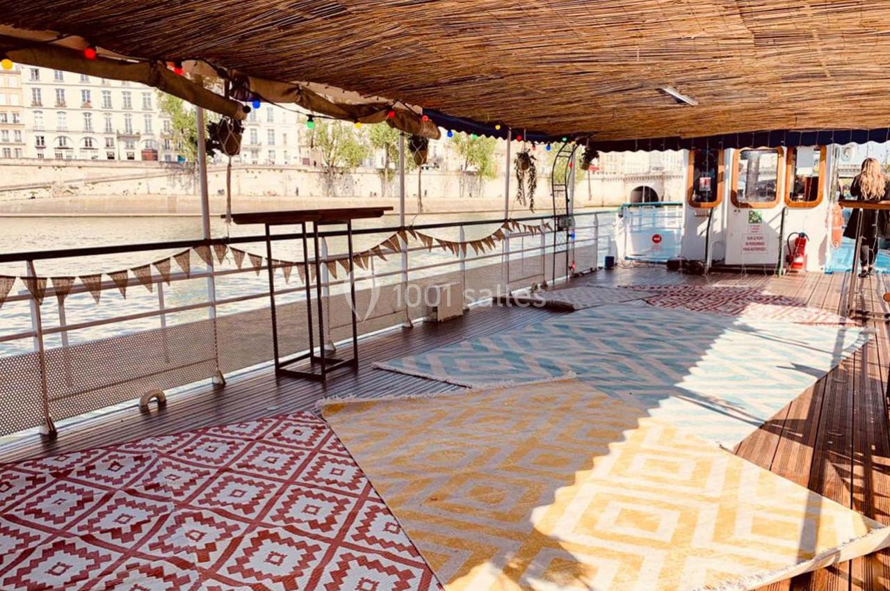 Pont d'une péniche décoré de tapis colorés et guirlandes, avec vue sur des bâtiments en bord de rivière.