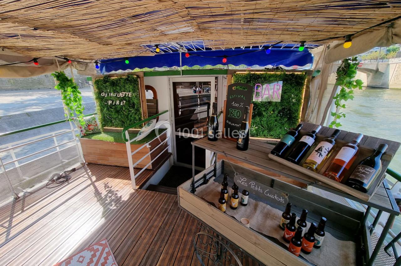 Terrasse d'une péniche aménagée avec un bar, des étagères de bouteilles de vin et une décoration végétale.