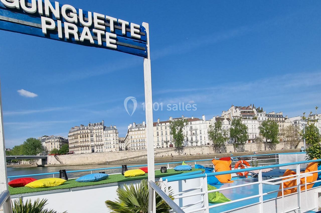 Terrasse colorée d'une guinguette flottante avec vue sur des immeubles et la Seine sous un ciel bleu.
