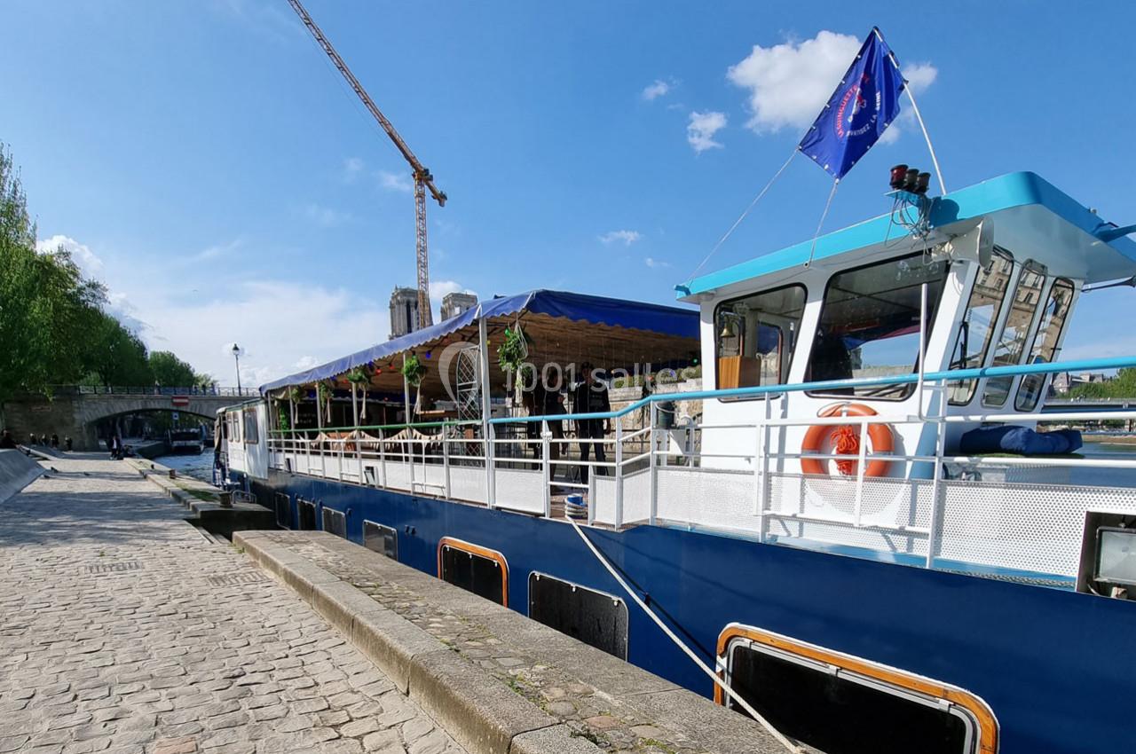 Bateau amarré le long d'un quai pavé, avec une terrasse couverte et une grue visible en arrière-plan.