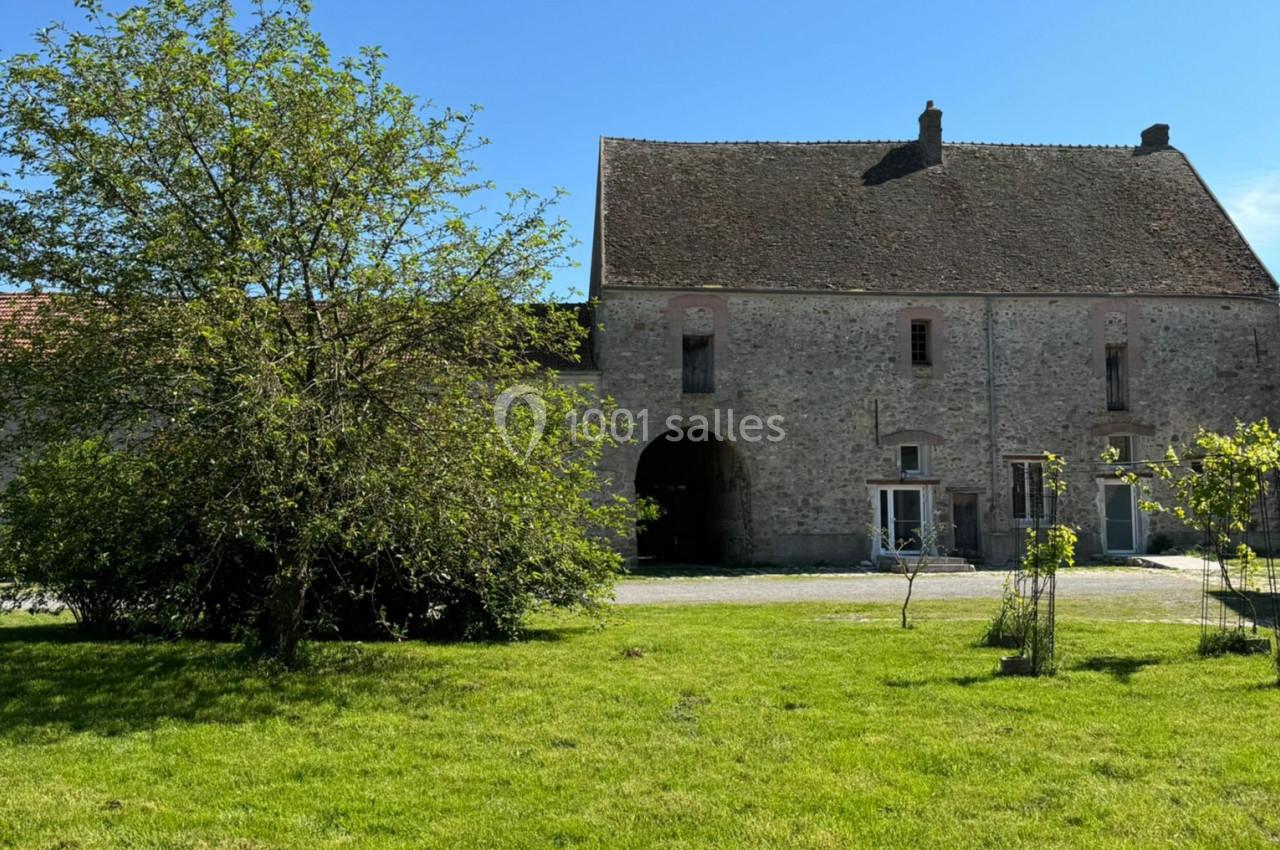 Bâtiment en pierre avec toit en pente, grande arche centrale, arbre et pelouse au premier plan sous un ciel bleu.