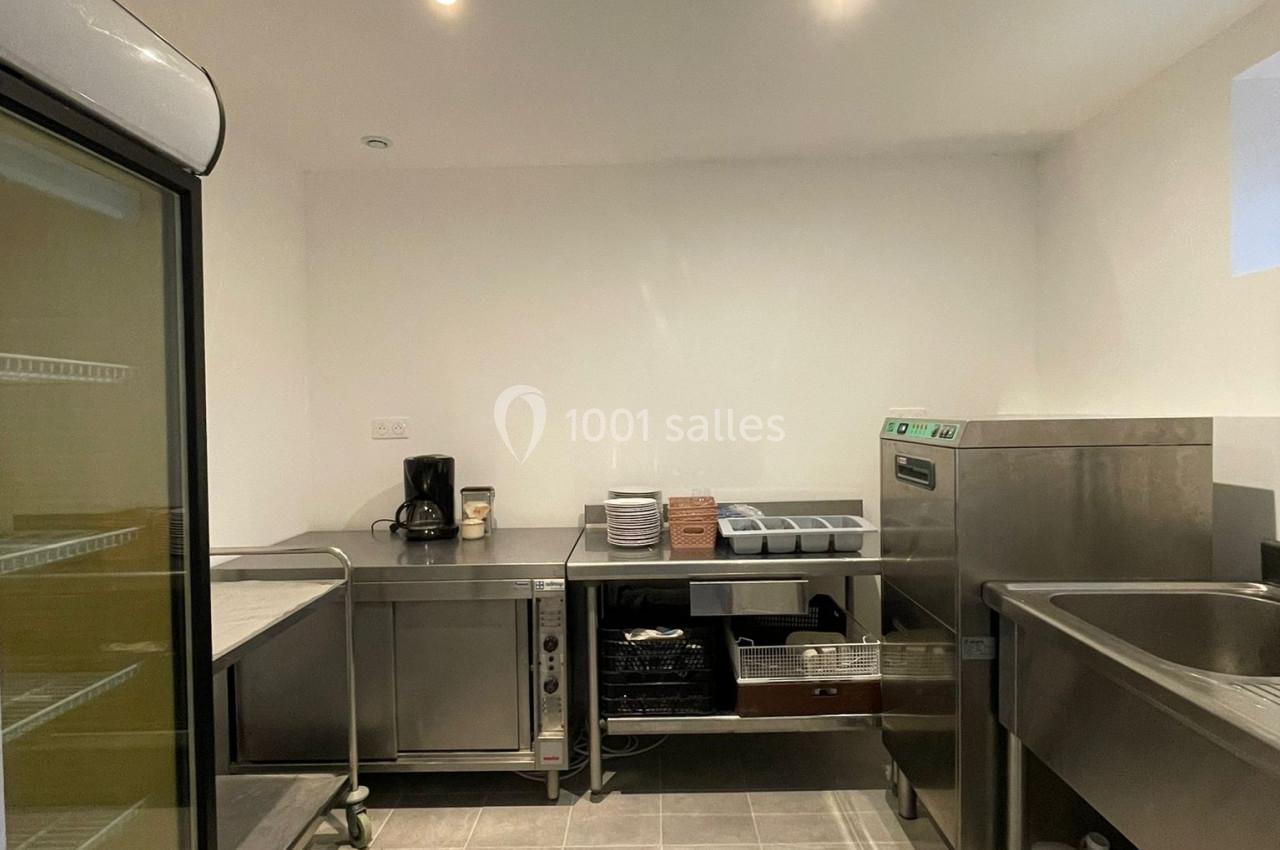 Cuisine professionnelle avec équipements en inox, évier, réfrigérateur vitré et plans de travail sous un éclairage lumineux.