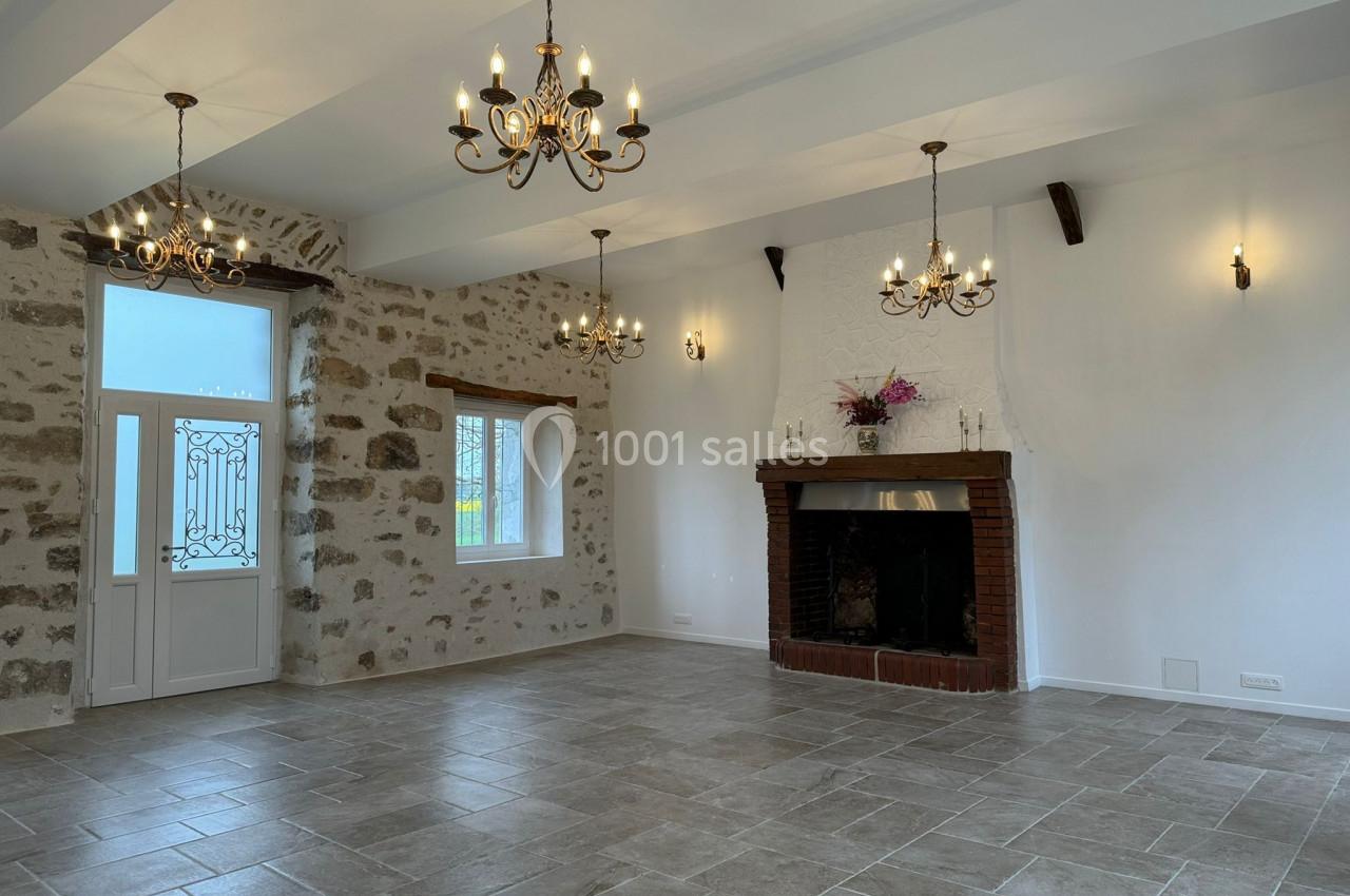 Salle lumineuse avec murs en pierre apparente, cheminée en briques, lustres suspendus et sol en carrelage gris.