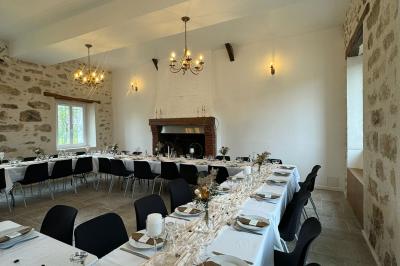 Salle de réception lumineuse avec tables en U décorées de nappes blanches, fleurs et bougies, murs en pierre apparente.