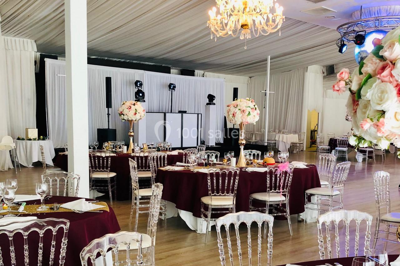 Salle de réception élégante avec tables dressées, nappes bordeaux, chaises transparentes et décorations florales.