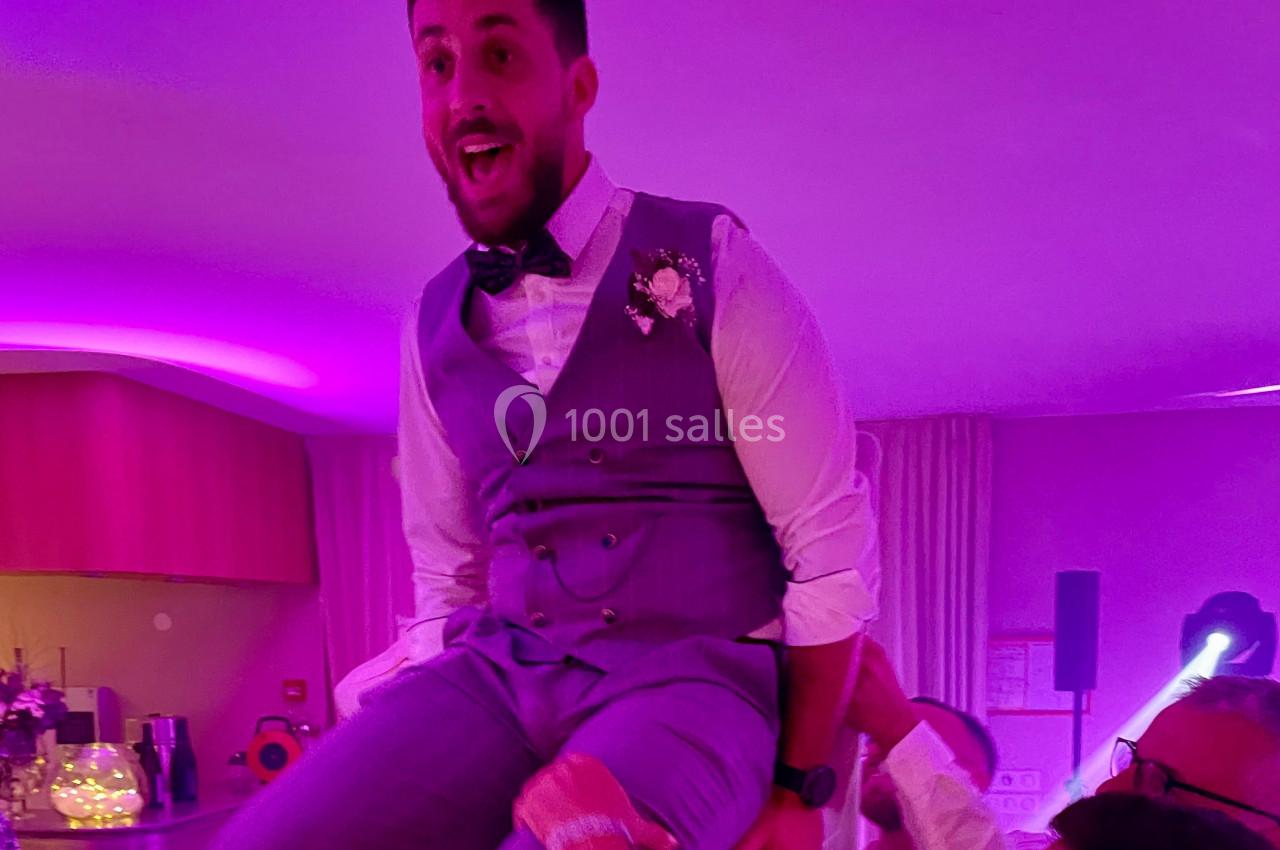 Un homme en costume gris, souriant, est porté sur les épaules de quelqu'un dans une ambiance festive éclairée en violet.