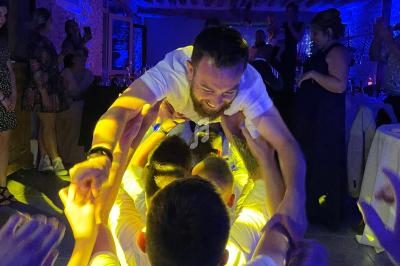 Un homme en chemise blanche est porté par un groupe de personnes lors d'une fête dans une ambiance lumineuse colorée.
