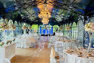 Salle de réception élégante avec tables décorées, chaises transparentes et lustres suspendus sous une verrière.