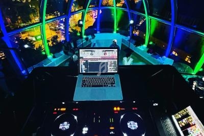 Poste de DJ avec platines et ordinateur dans une salle vitrée illuminée de lumières colorées, vue nocturne en arrière-plan.