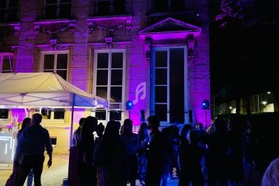 Groupe de personnes rassemblées devant un bâtiment illuminé de lumières violettes, avec une tente blanche à gauche.