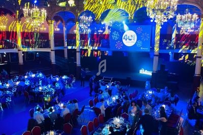Salle de réception illuminée avec des tables dressées, des invités assis et une scène avec écran projetant un logo.