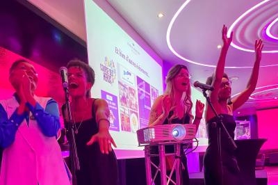 Quatre personnes chantent sur scène avec des micros, un projecteur et un écran en arrière-plan dans une salle éclairée.