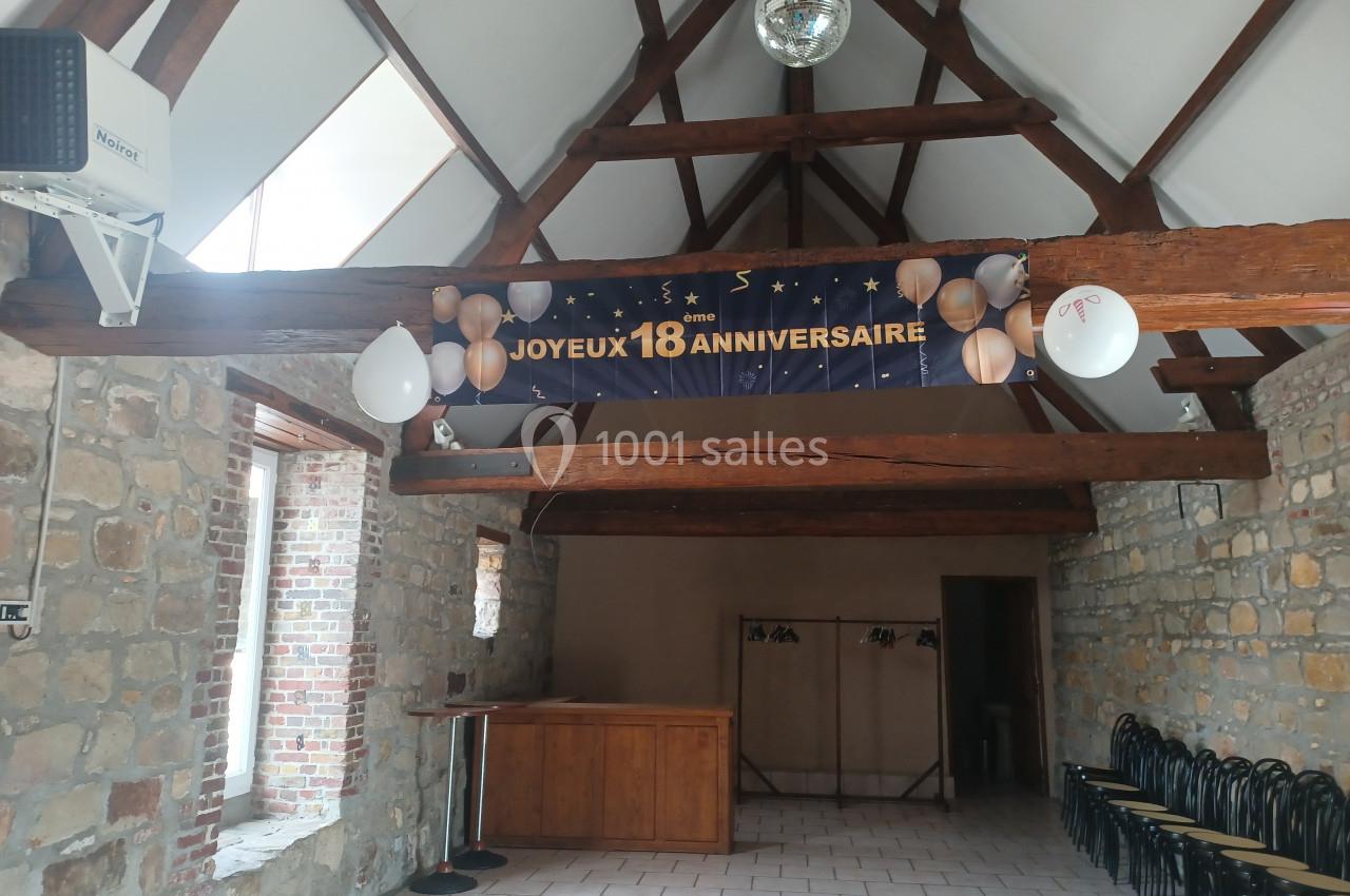 Salle en pierre avec poutres apparentes, décorée d'une banderole ’Joyeux 18ème anniversaire’ et de ballons blancs.