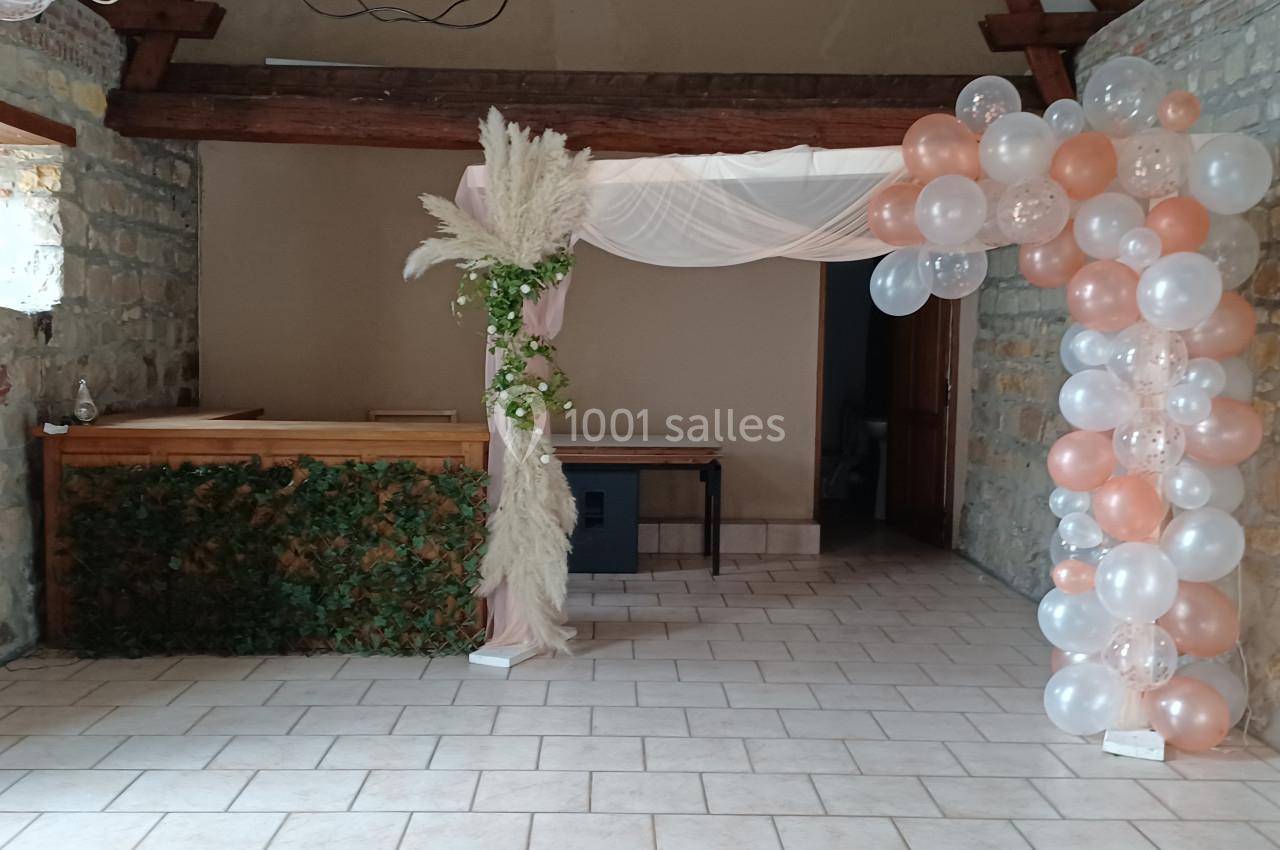 Décoration de salle avec arche florale, ballons blancs et roses, et comptoir en bois orné de feuillage.