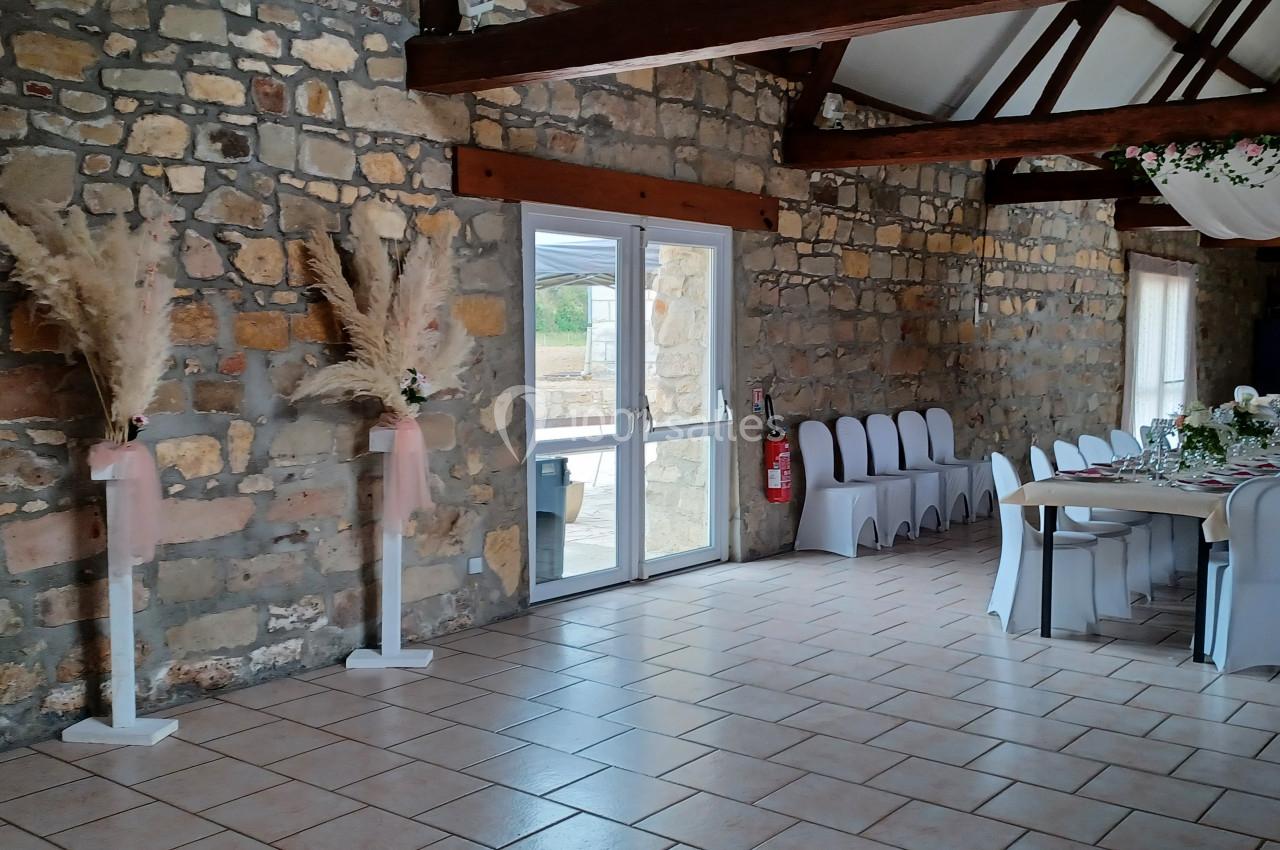 Salle en pierre avec poutres apparentes, décorée pour un événement, tables dressées et chaises blanches alignées.