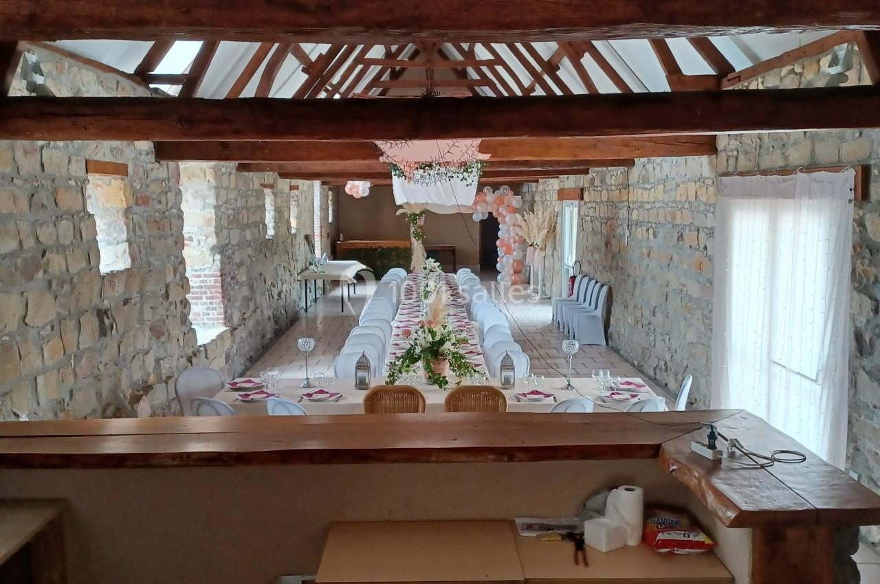 Salle en pierre avec poutres apparentes, tables décorées pour un événement, alignées sous une guirlande florale suspendue.