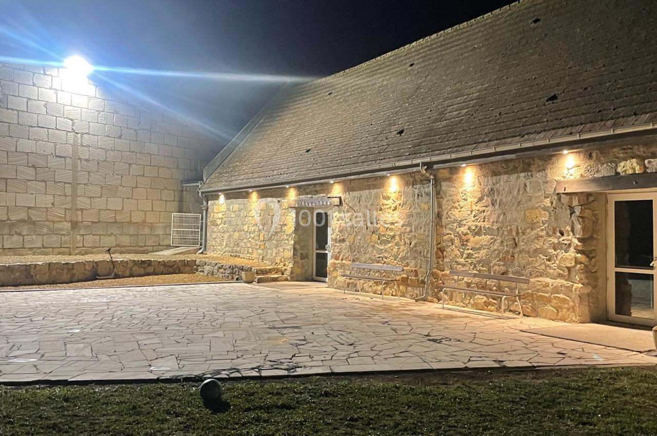 Cour en pierre éclairée la nuit, bordée par un bâtiment en pierre avec des lumières murales et un espace gazonné au premier…