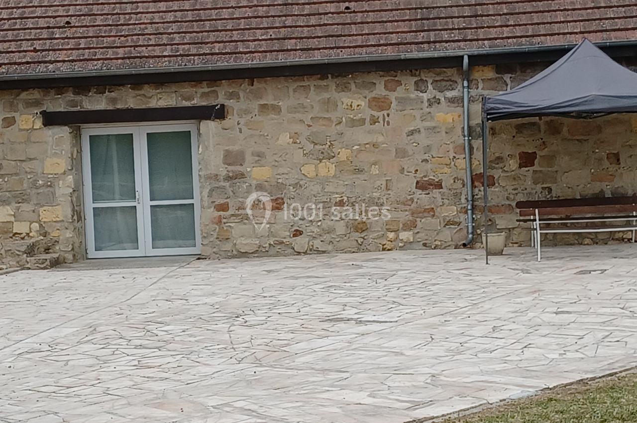 Mur en pierre avec une porte vitrée, une terrasse pavée, un banc en bois et un petit abri gris.