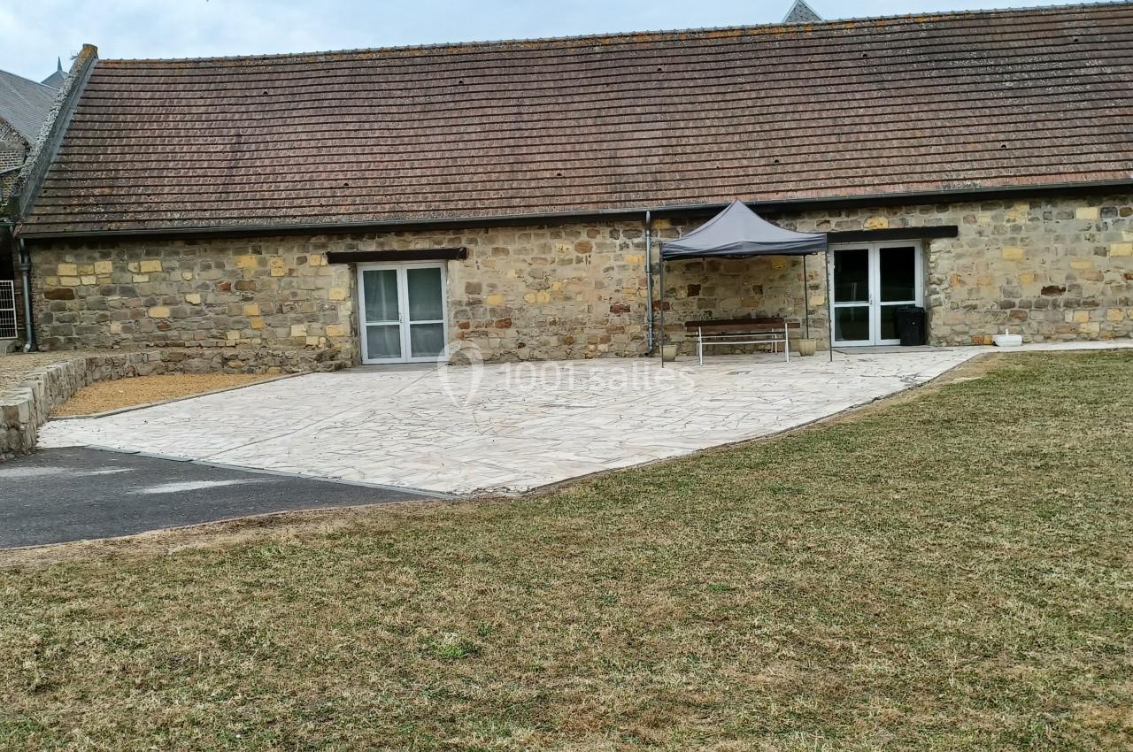 Bâtiment en pierre avec toit en tuiles, terrasse pavée, petite tente et pelouse au premier plan.