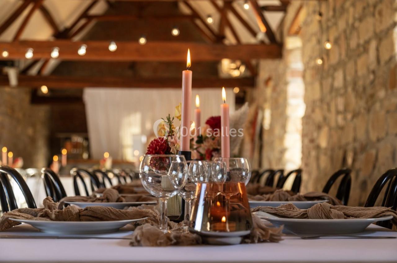 Table décorée avec des bougies allumées, des fleurs et des verres, dans une salle en pierre avec poutres apparentes.