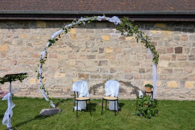 Arche décorée de feuillage et de tissu blanc avec deux chaises recouvertes, installées sur une pelouse devant un mur en…