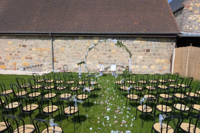 Arche décorée de feuillage et de tissu blanc avec deux chaises recouvertes, installées sur une pelouse devant un mur en…