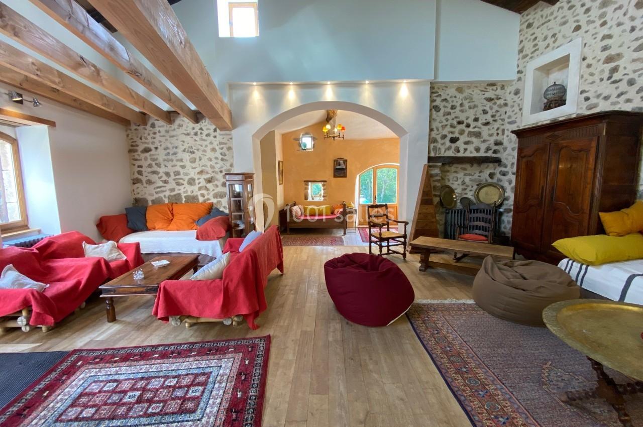 Salon spacieux avec murs en pierre, poutres apparentes, canapés colorés, tapis et lumière naturelle.