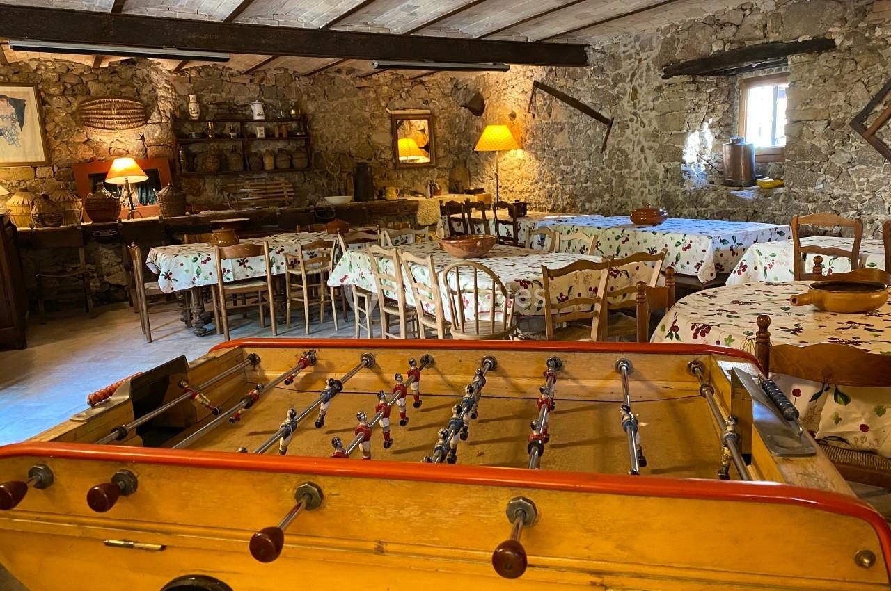 Salle rustique avec murs en pierre, tables recouvertes de nappes colorées et un baby-foot au premier plan.